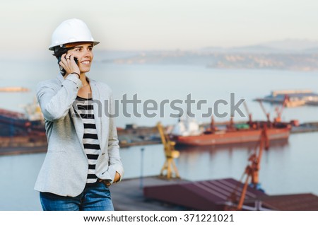 successful independent engineer woman talking on the phone and smiling at industrial harbor with safety helmet. Pioneer woman at work. Safety at work.