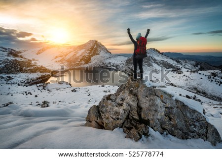 Hiker woman with backpack and hat rising arms in victory sign on snowy mountain peak at Covadonga lake, beautiful snow winter landscape sunset scene on a touristic location of Asturias, Spain, Europe.