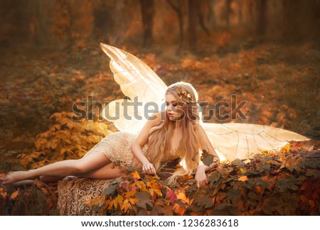 slim girl became a fairy, a model with blond long hair and golden wreath on leaves in the forest in a beige long dress with bare legs, has glowing wings behind her back, atmospheric autumn art photo.