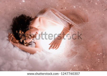 gentle image of angel sleeping next to the moon in pink light clouds, naked body of slim girl lying in a mist, covered with glitter sequin, princess of the night with dark hair and a wonderful wreath