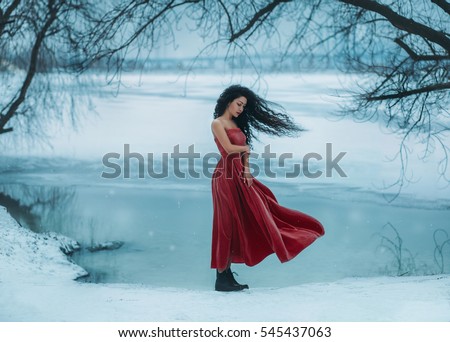 Beautiful brunette girl badly frozen. Background shore of a frozen river, water and ice. She is dressed in a beautiful red dress. The wind waves are long, curly hair. Fantasy photo, creative color.