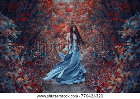 Mysterious sorceress in a beautiful blue dress. Her hair and dress are fluttering in the wind. Background bright, autumn, fiery forest with cold tones. Artistic Photography