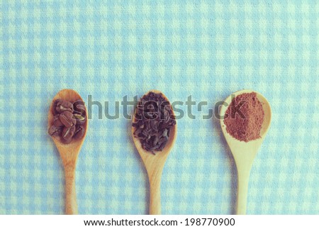 Roasted coffee beans, tea, cocoa powder in wooden tea spoons on a table with pastel tablecloth, vintage filter