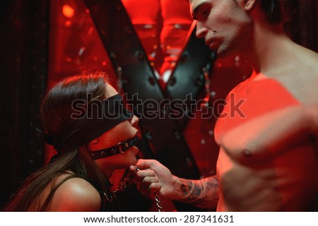 BDSM bondage scene. Girl with gag  withheld by neck chain by an ideal man in red room.