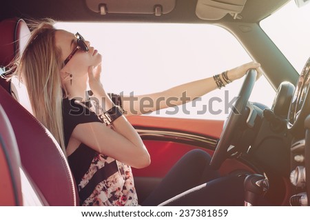 Blondie young girl at the wheel of sport car with red interior with black sunglasses and leather armlets seating sideward with hand near her check and wind in her hair and looking at the road