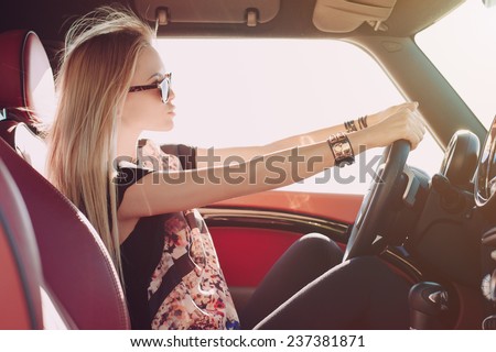 Blondie young girl at the wheel of sport car with red interior with black sunglasses and leather armlets with metal inserts, seating sideward with two hands on the wheel and looking at the road