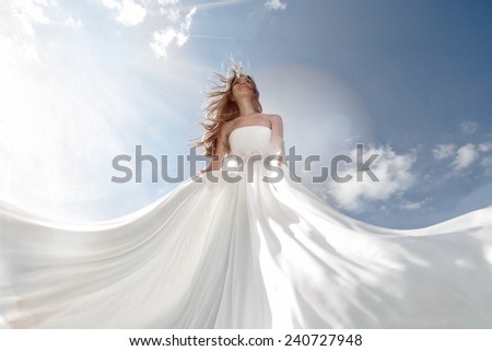 Young women in white floor-length dress with long lower hem waved in the breez standing in the middle and looking up with light blue sky with a lot of clouds behind her