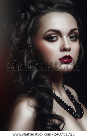 Gothic portrait of young green-eyed woman with black curles, black necklace black wings and brown smoky eyes and bardic lipstick standing half turned to the right and looking at you black background