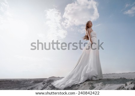 Young women in white wedding floor-length dress with long lower hem waved  in looking up with light blue sky with a lot of clouds behind her