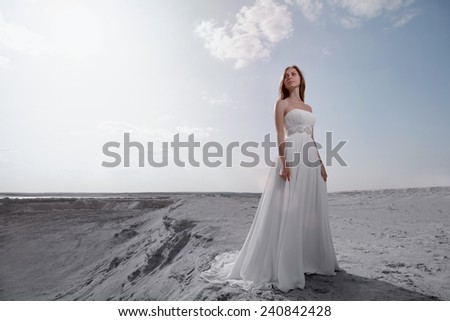 Young women in white wedding dress with long lower hem waved  in looking away with light blue sky with a lot of clouds behind her
