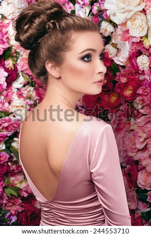 Soft facial portrait of young charming woman in rosy skin-tight gown with V-shaped cut and open back, dark smoky eyes and natural lipstick standing behind on flowered background made of peony