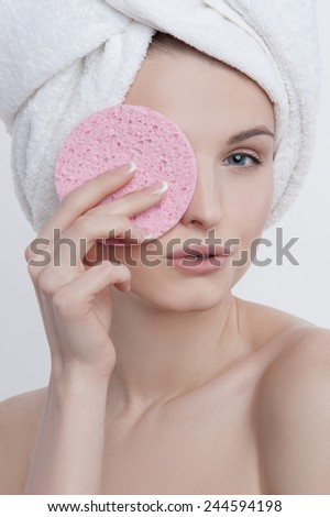 Portrait of good-looking blue-eyed young lady with natural make-up, french manicure and clear skin with white towel on her head and pink rough sponge in her left hand closing her left eye.