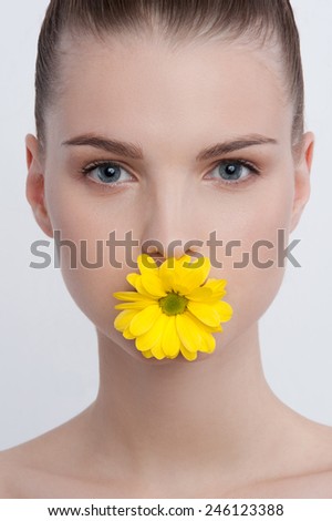 Facial simple portrait of cute young girl with blue eyes, natural make-up and pony tail, with yellow small chrysanthemum in her lips looking at you on white background