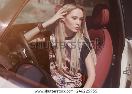 Soft portrait of blondie young girl at the wheel of sport car with red interior, black leather armlets her arm, long strighten hair, natural makeup looking to the right