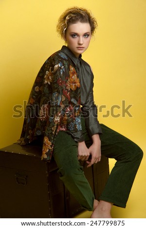 Fashion portrait of woman with natural make up. Wears casual grey blouse, moss green velvet trousers and dark jacket with big flowers. No boots. Seats on old chest on yellow background.
