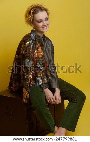 Fashion portrait of young woman with natural make up. Wears casual grey blouse, moss green velvet trousers and dark jacket with big flowers. No boots. Seats on old chest on yellow background.