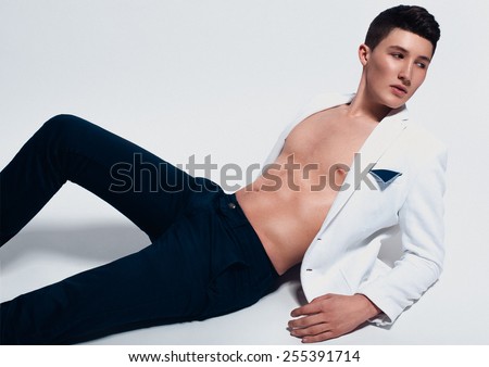 Fashion portrait of handsome young man in dark jeans and white jacket laying on the floor and looking to the left on white background