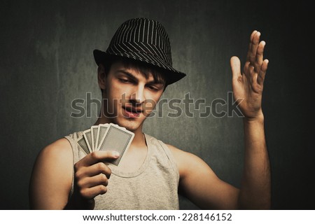 annoyed poker player hold cards in one hand and gesturing with the other hand, wearing hat and gray tank top, studio shot