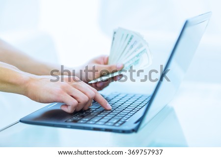 Close-up of hands shopping/paying online using laptop while holding US dollars..