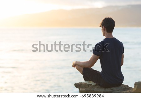 Young man meditating on top ocean cliff during sunset.