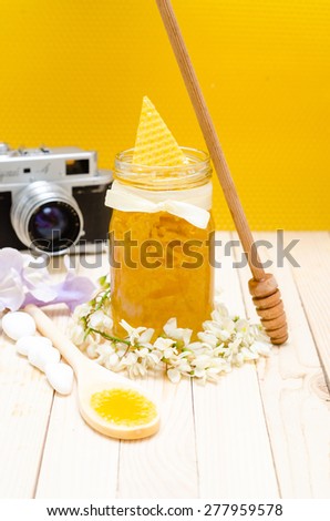 Locust Honey Retro Jar with locust blossoms, and small white stones on retro wooden table with Honeycomb and camera in the background. Selective Focus on glass of jar