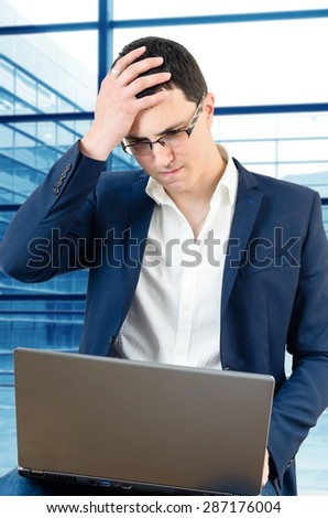 Young businessman wearing glasses and holding his head on the airport with laptop in his lap