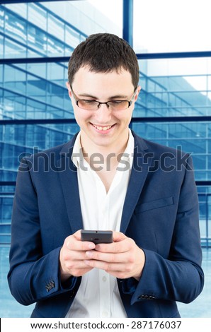 Young business man wearing glasses and texting a massage on his smartphone over big office\'s windows background