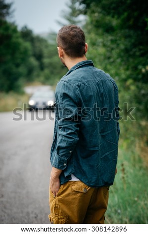 Young man standing on the road, looking in the car coming towards him. Vintage Instagram style effect, soft and selective focus, shallow DOF, low light, grain texture visible on maximum size