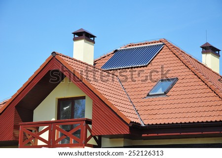 Solar water panel heating on red tiled house roof with lightning protection and chimney against blue sky