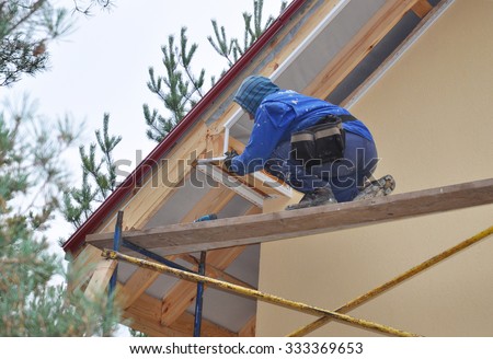Roofer on the Corner of House  Install Soffit. Roofing Construction. Soffit and Fascia is Usually Constructed of Vinyl, Wood or Aluminum and is Installed on the Underside of Roof Overhangs and Eaves.