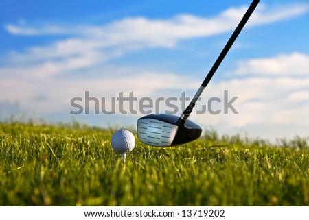 Golf ball about to be struck by driver with grass and blue sky.