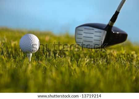 Golf ball about to be struck by driver with grass and blue sky.