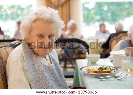 An smiling elderly woman sits down to enjoy a meal at her nursing home care center with a sunny background.