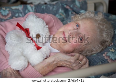 An smiling elderly woman lies down to enjoy a rest at her nursing home care center on a sunny day holding a bear.