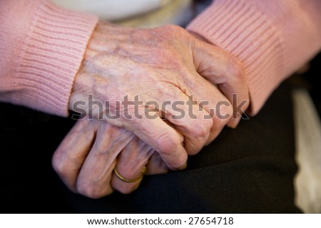 A elderly woman crosses her hands politely, wearing a pink sweater in her nursing home care center.