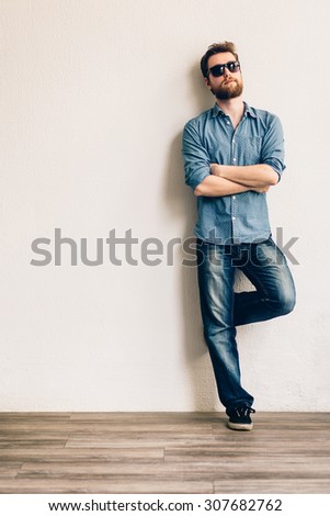 Young handsome man with sunglasses, leaning against a white wall. Hands crossed.