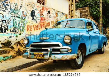 HAVANA, CUBA - DECEMBER 2, 2013: Old broken American blue car parked in the old town of Havana, next to wall paintings presenting remnants of the Cuban Communist Revolution