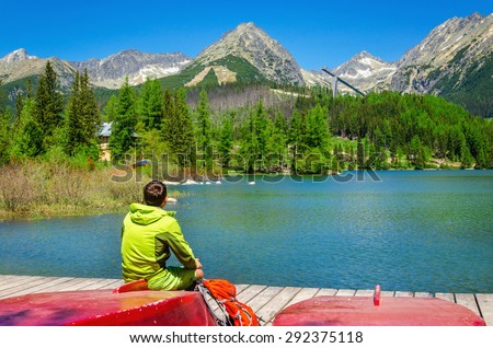 Young man sitting on red boat at the pier mountain lake in the background of the high peaks of the mountains, Strbske Pleso, Slovakia