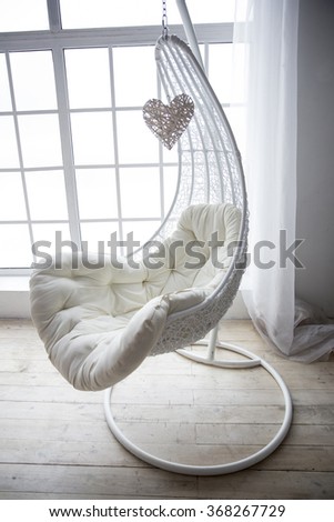 beautiful white hanging chair with a gentle soft mattress in the form of drop on the wooden floor in the corner by the window with a transparent curtain