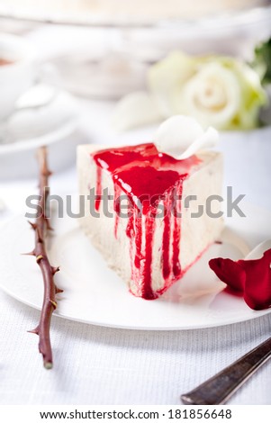 Cheesecake, mousse cake slice with red berry topping and rose petals on a white plate with a rose stem with thorns