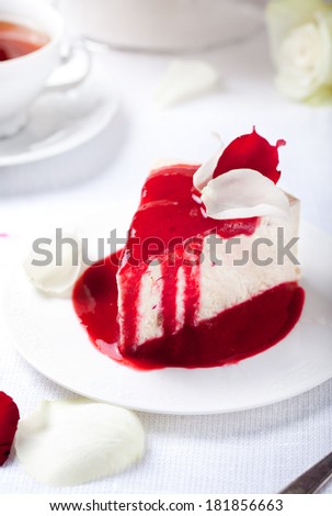 Cheesecake, mousse cake slice with red berry topping and rose petals on a white plate with a rose stem with thorns