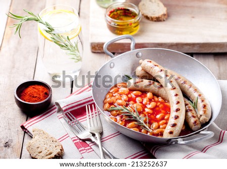 Grilled sausages with beans in tomato sauce in with rosemary and paprika in a vintage pan on a wooden background.