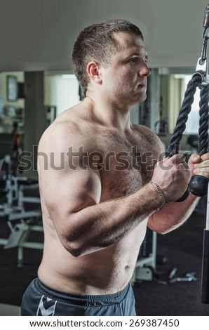 Young man at the gym exercise on triceps extension arms standing in the simulator with a rope. Toning, hard light.