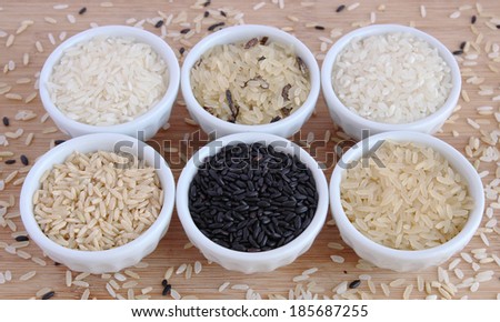 Six varieties of uncooked rice in white bowls on wooden board