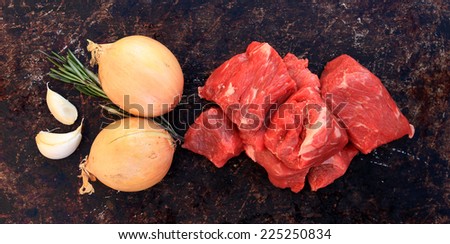 Raw beef cubes with onion, garlic and rosemary on brown rustic background