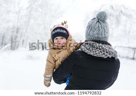 A boy son looks out from behind mother. The mother carries the baby in winter park. Closeup portrait