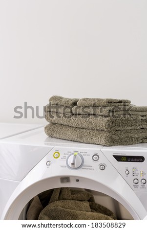 Several folded towels on top of an electric clothes dryer in a laundry room. Other towels still in dryer are visible through the opened door.
