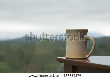 Rainy, foggy mountains in the background with a coffee mug on the corner of a table near a window.