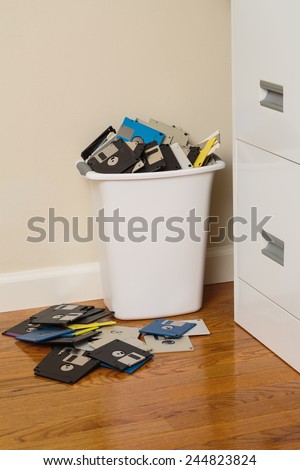 Wastebasket overflowing with 3.5-inch floppy disks beside a file cabinet.