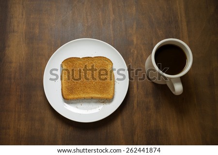 Slice of plain dry whole wheat toast on a white plate and cup of coffee on a rustic table shot from directly above.